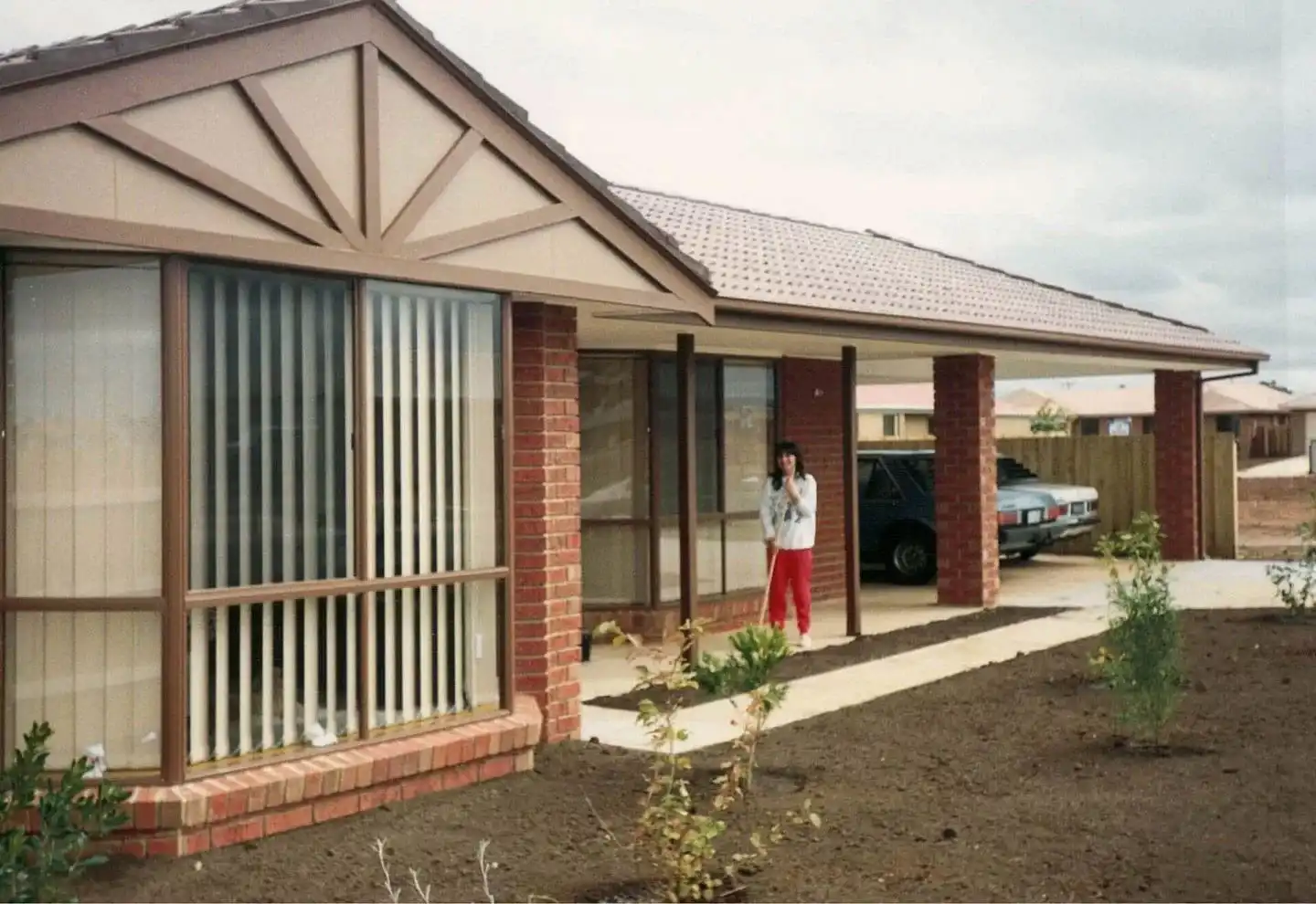 A photograph of a woman standing next to a single storey house with a front yard and a garage space.