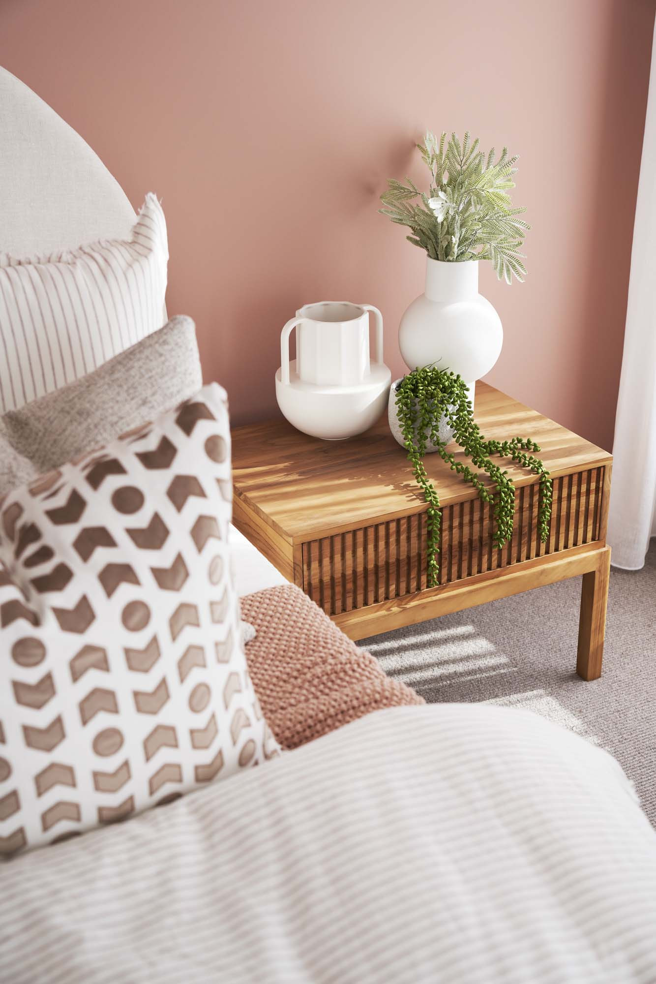 Close up photo that shows the back right edge of a bed and a wooden table beside the bed. Behind it is a peach coloured feature wall, all part of the Simonds Wallaby 26 home design